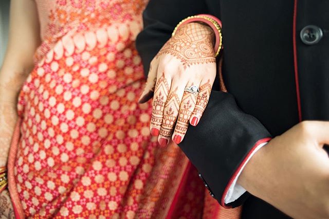 A bride's hand adorned with mehndi and a wedding ring rests on the groom's arm during their wedding ceremony.