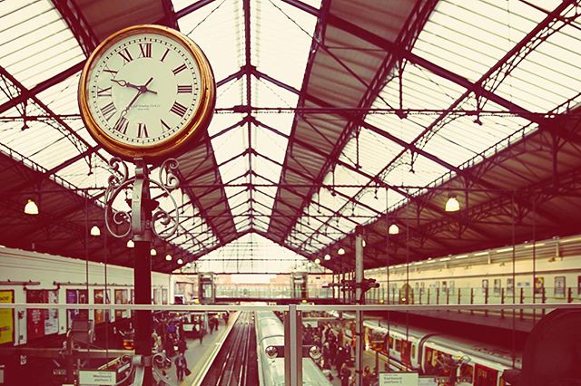 A clock tower stands inside a bustling train station with platforms and people waiting. 