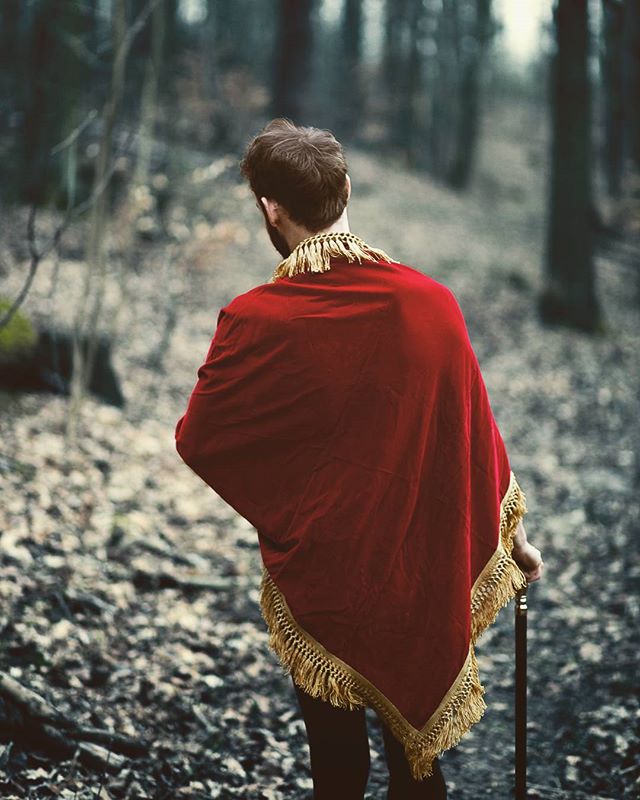 A man in a red cape walks through a forest, holding a walking stick. The image has a vintage and mysterious feel.