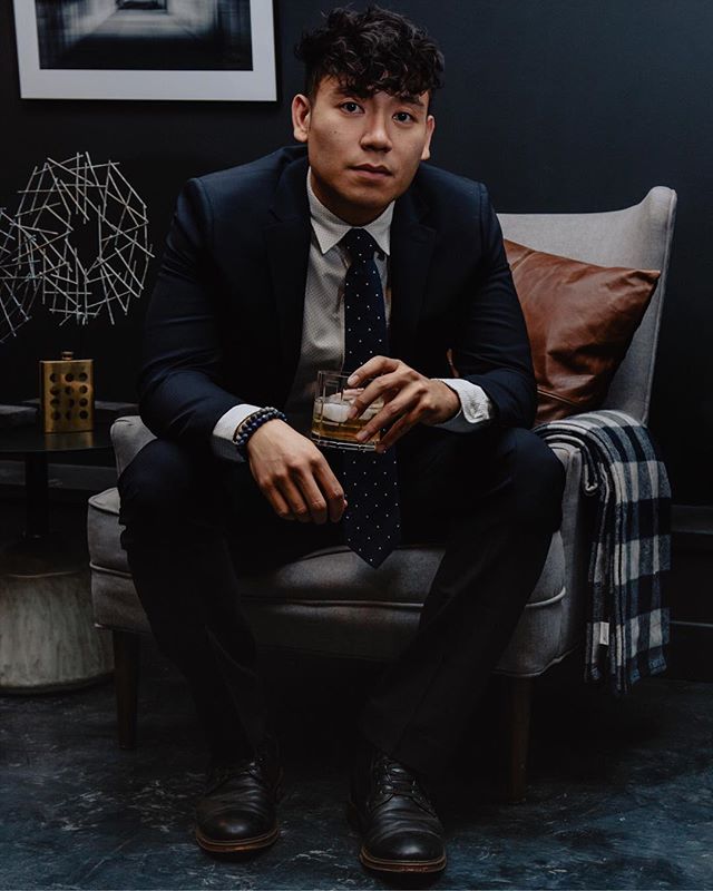 A stylish man sits in a chair, holding a glass of whiskey in a sophisticated indoor setting.
