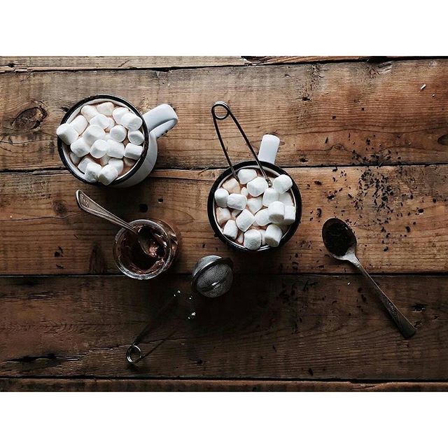 Overhead view of mugs with hot chocolate, marshmallows, and a spoon on a wooden table.