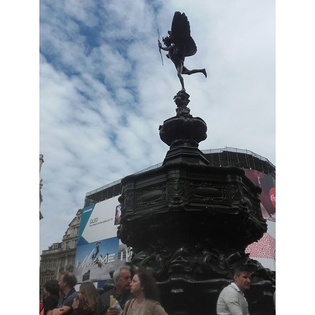 The statue of Eros stands tall in London's Piccadilly Circus, surrounded by people under a cloudy sky.