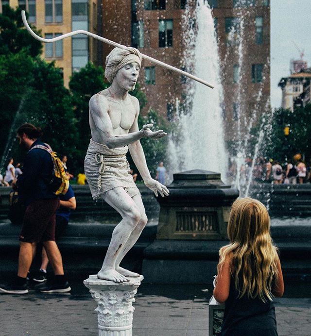 A street performer entertains a young girl near a fountain in a bustling city park.