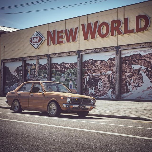 A vintage brown Toyota car parked in front of a New World store with a mural on the building.