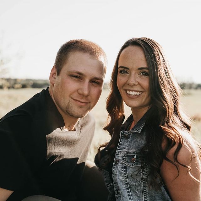 A smiling couple poses outdoors in a sunny field, creating a natural and cheerful portrait.
