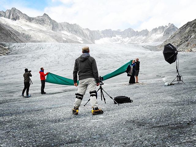 Outdoor photo shoot on a glacier with a couple posing and crew assisting in front of mountains.