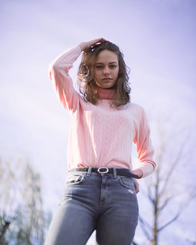 A young woman poses outdoors in a pink sweater and jeans, looking confidently at the camera.