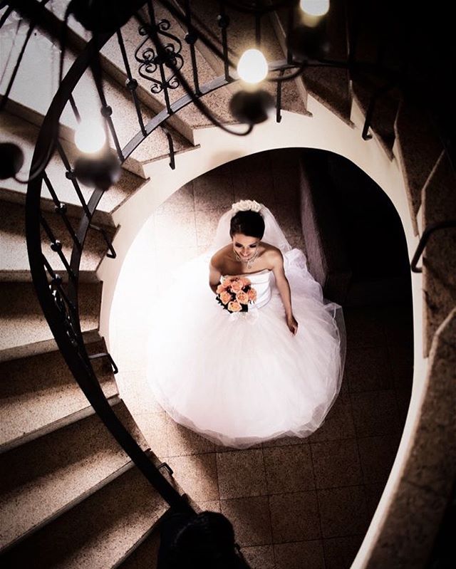 A bride in a white gown descends a spiral staircase, holding a bouquet for her wedding day.