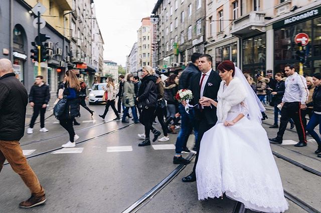 Bride and groom crossing a city street with wedding party and pedestrians in background.
