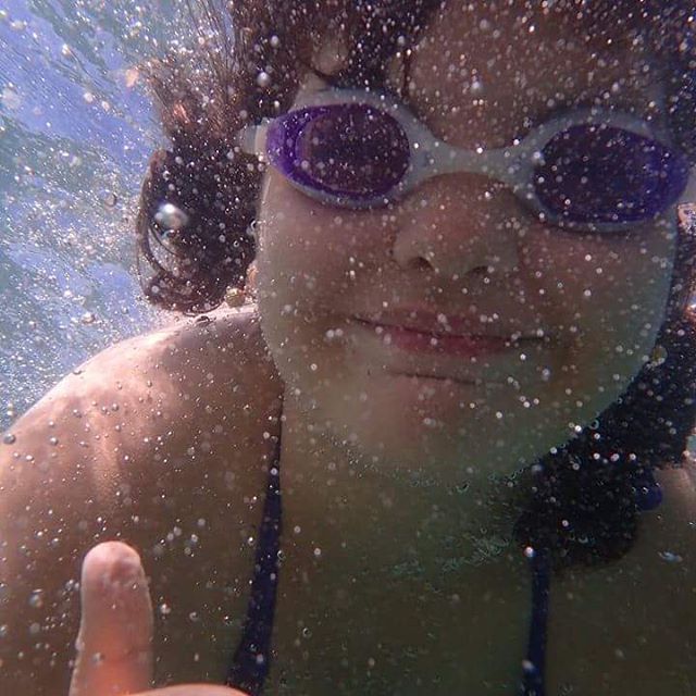 A cheerful person swimming underwater with goggles gives a thumbs-up, capturing a fun and playful moment.