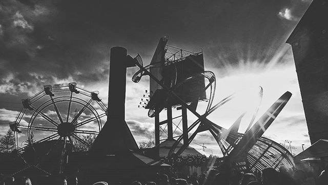 A black and white shot of a fairground with a ferris wheel and roller coaster in the background, and a crowd in the foreground.