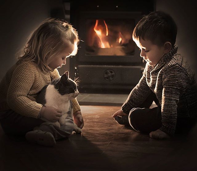 Two children sit quietly with their cat near a warm fireplace, creating a cozy, intimate atmosphere.