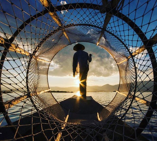 A lone fisherman stands in his boat at sunset, framed by a traditional fishing net on a serene lake.