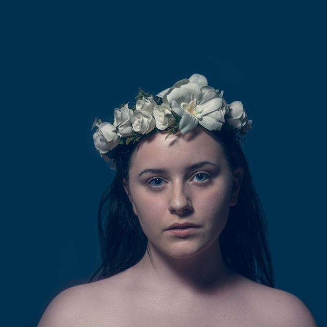 A young woman with a floral crown poses against a blue background in a dreamy and ethereal portrait.