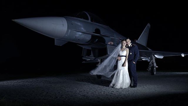 A bride and groom stand in front of a fighter jet on their wedding day, celebrating their union.