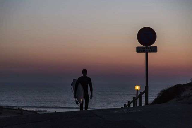 A lone surfer walks on the beach at dusk with a surfboard under his arm, silhouetted against the colorful sky.