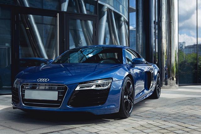 A blue Audi R8 sports car is parked outside a modern building, showcasing its sleek design and urban appeal.