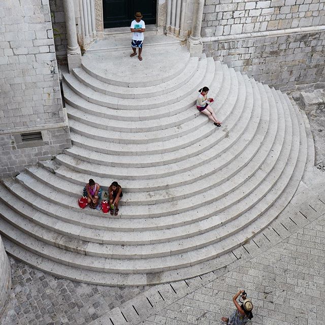 People sit and stand on the curved stone stairs of a historic building in a European city, enjoying the sights.