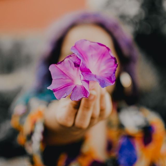 A woman with purple hair holds up vibrant pink and purple flowers, creating a dreamy, soft, and editorial feel.