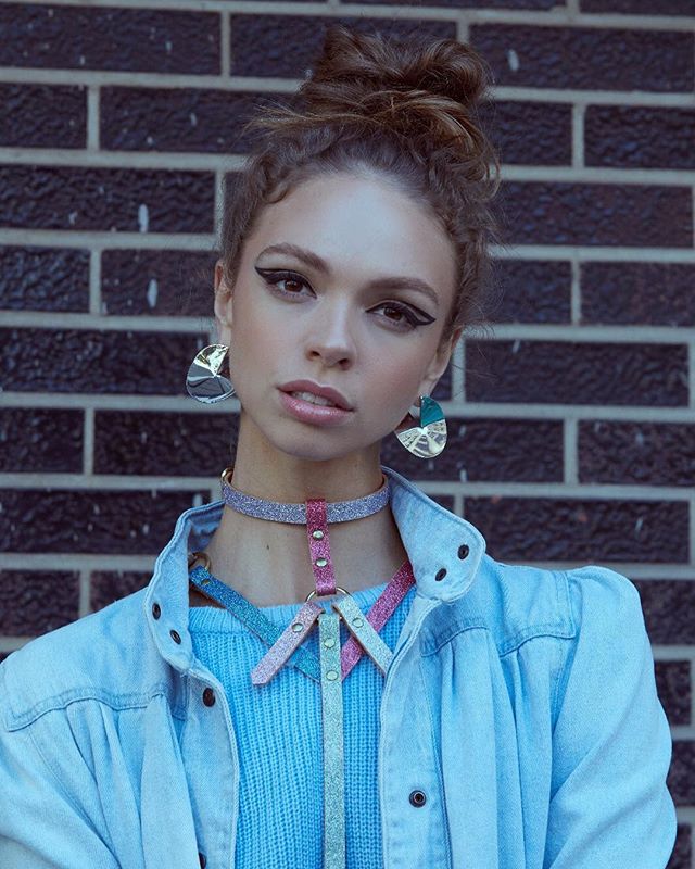 A young woman with stylish makeup poses against a brick wall, showcasing modern jewelry and fashion.