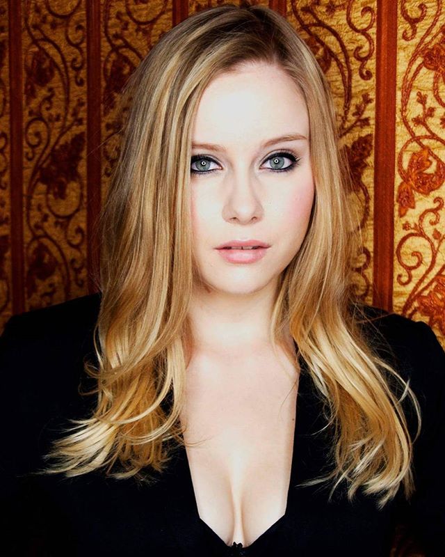 A blonde woman poses in a medium shot with soft lighting against an ornate background.