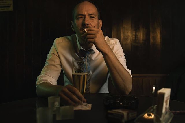 A man in a white shirt is sitting at a table with a beer and a cigarette.