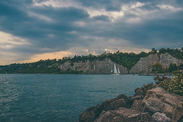 A scenic view of a sailboat sailing along the cliffs in a calm sea under an overcast sky.