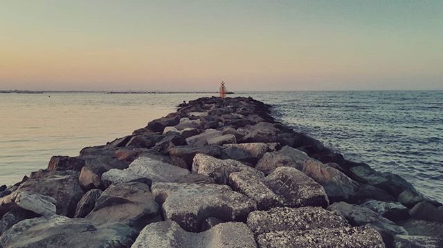 A rocky jetty stretches into the calm sea under a serene sky, featuring a beacon at the end.