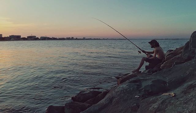 A man fishing off the rocks at sunset near the ocean, enjoying outdoor recreation near the city.