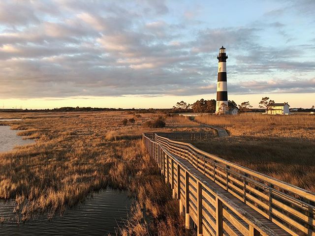 A scenic view of a black and white lighthouse on the coast with a wooden boardwalk leading towards it.