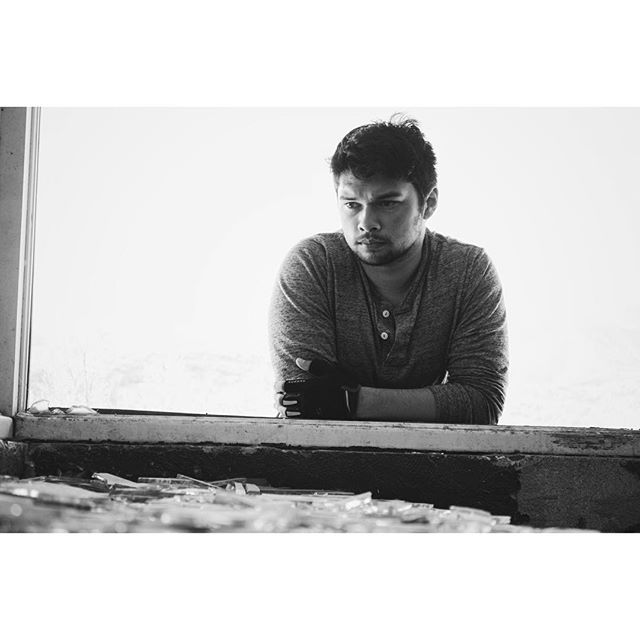 A pensive man gazes into the distance in a black and white portrait, standing by a window in a muted emotional state.