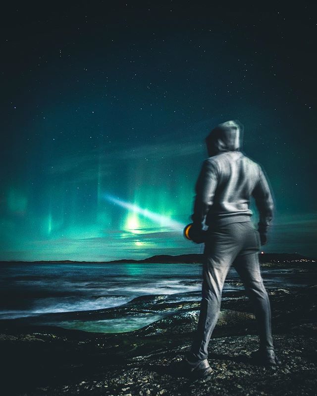 A lone man stands on the rocky coast gazing at the mesmerizing aurora borealis in the night sky.