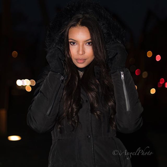 A woman with long dark hair poses in a black hooded coat at night in an urban setting.