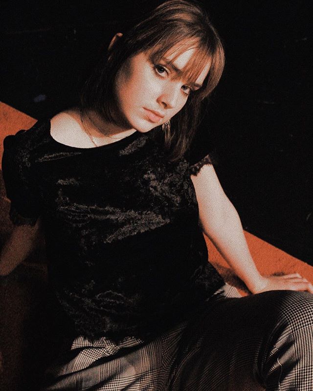 A woman poses in a dimly lit studio with a thoughtful expression, showcasing fashion and lifestyle aesthetics.