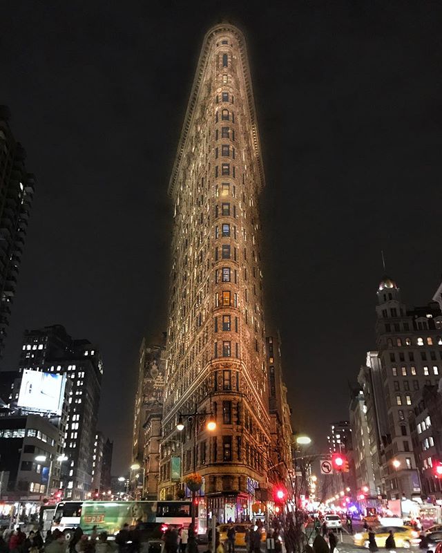 The iconic Flatiron Building glows at night in New York City, surrounded by bustling city life.
