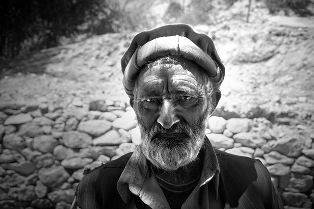 A striking black and white portrait of an elderly man with a weathered face in front of a stone wall.