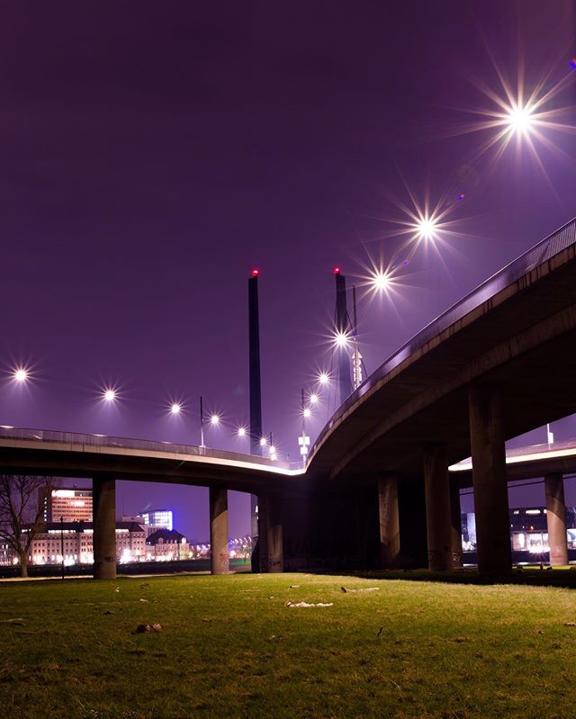 A night view of an elevated roadway and city buildings, brightly lit with streetlights, against a dark purple sky.