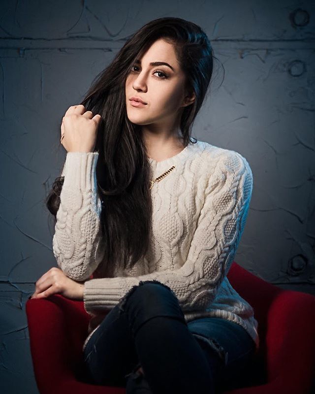 A young woman with long brunette hair poses in a white sweater and jeans, seated in a red chair.