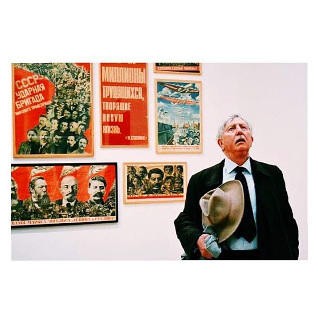 A man in a suit looks up at Soviet-era posters in an art gallery, holding his hat.
