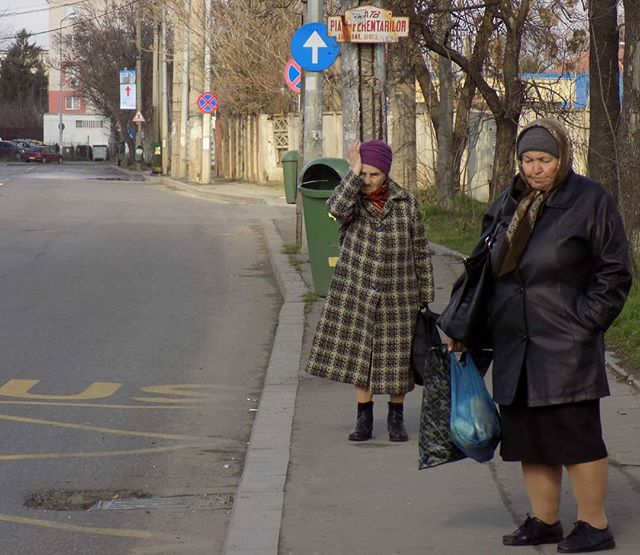 Two elderly women stand on a sidewalk on a city street, dressed in outerwear and carrying bags.