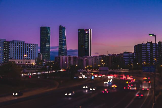 A cityscape with tall buildings and blurred lights on a highway under a vibrant purple and blue sky.