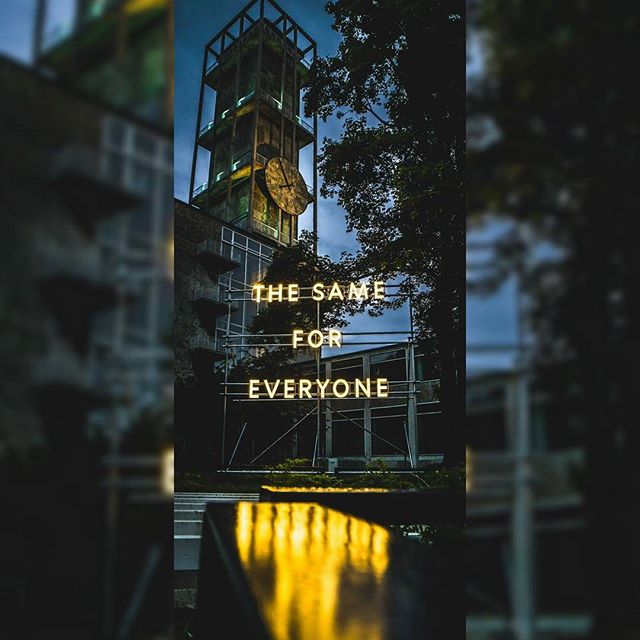 Illuminated signage 'The Same For Everyone' is displayed near a clock tower at night in an urban setting.