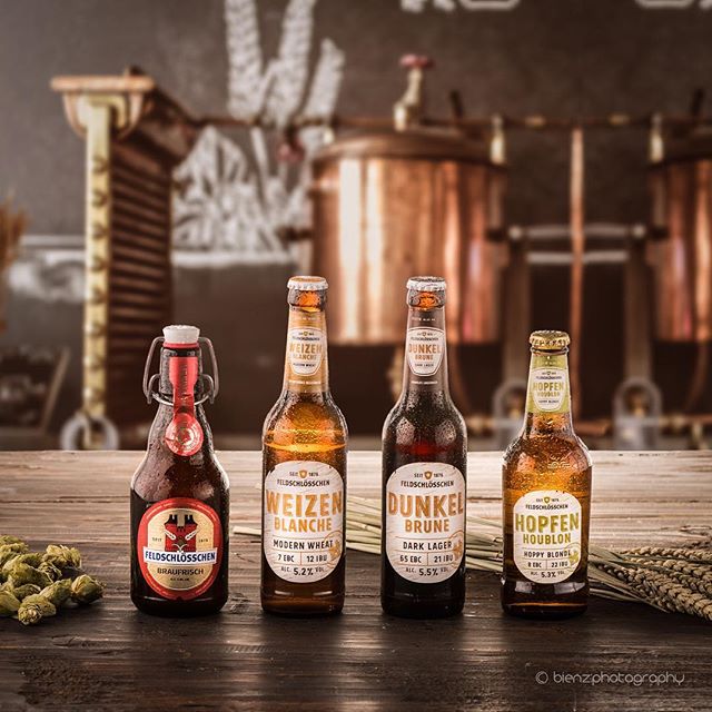 Four Feldshlosschen beer bottles stand side by side on a wooden surface in a studio setting.