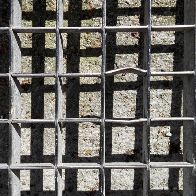 A close-up view of a gray metal grate creating an abstract grid pattern with visible shadows and texture.