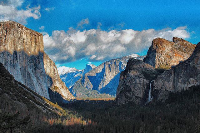 Scenic vista of a valley with mountains, a waterfall, and a forest under a blue sky with clouds.