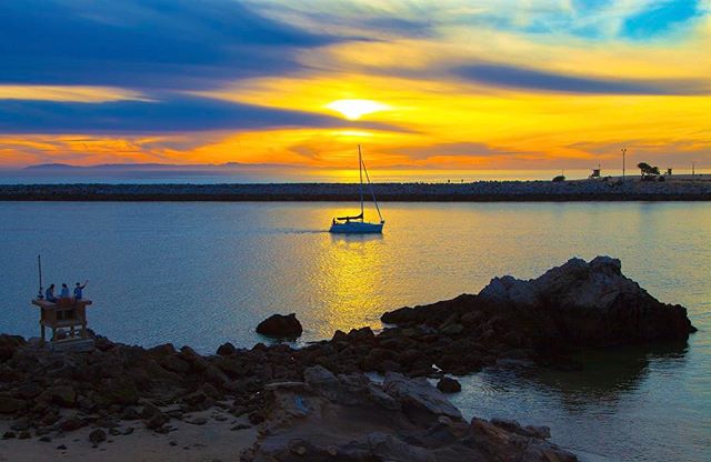 A sailboat is silhouetted against a vibrant sunset in a tranquil harbor with rocky coastlines. Ideal for travel or vacation campaigns.