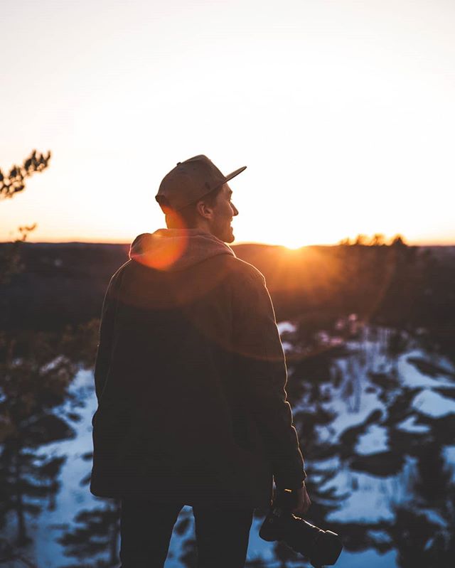 A lone photographer gazes at a serene sunset over a wintery landscape, capturing nature's beauty.