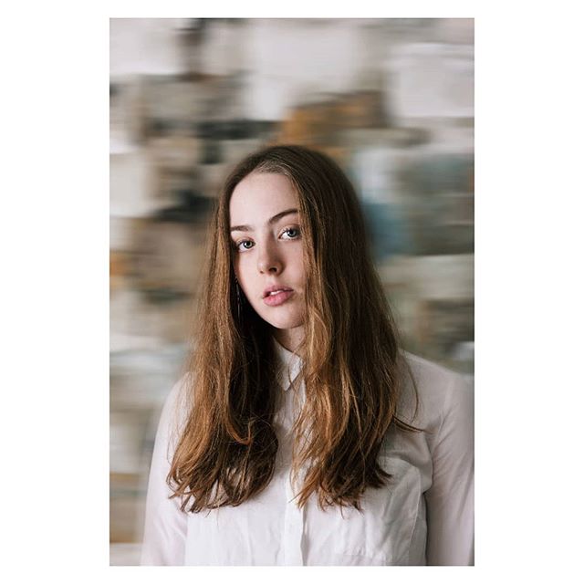 A young woman with long brown hair poses in front of a blurred neutral backdrop.