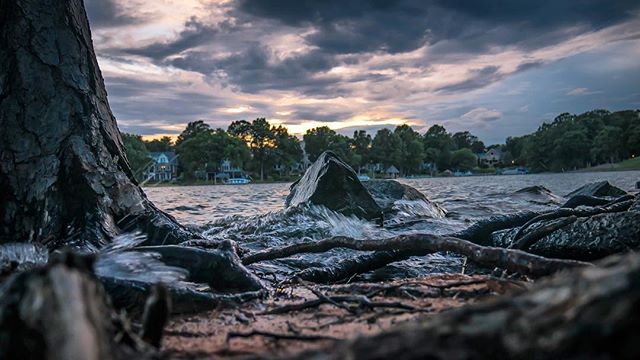 A serene view of a lake with trees, rocks, and moody clouds, captured from a low angle emphasizing the natural beauty.