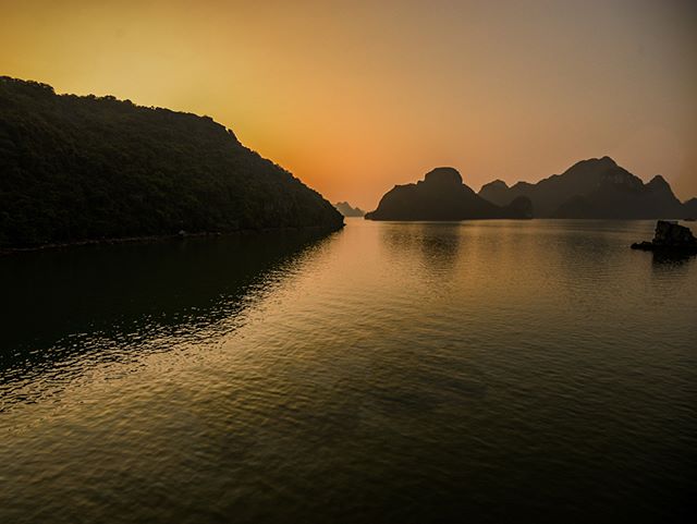 A tranquil seascape featuring islands silhouetted against a warm, orange-toned sky at sunset or sunrise.